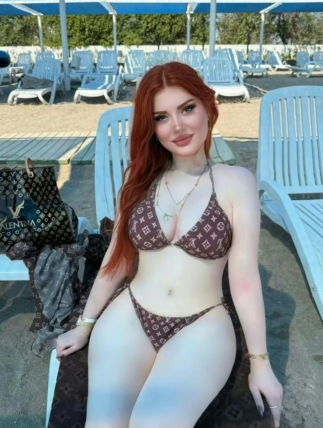 Woman in a brown LV-pattern bikini posing on a poolside chair area with blue lounge chairs in the background