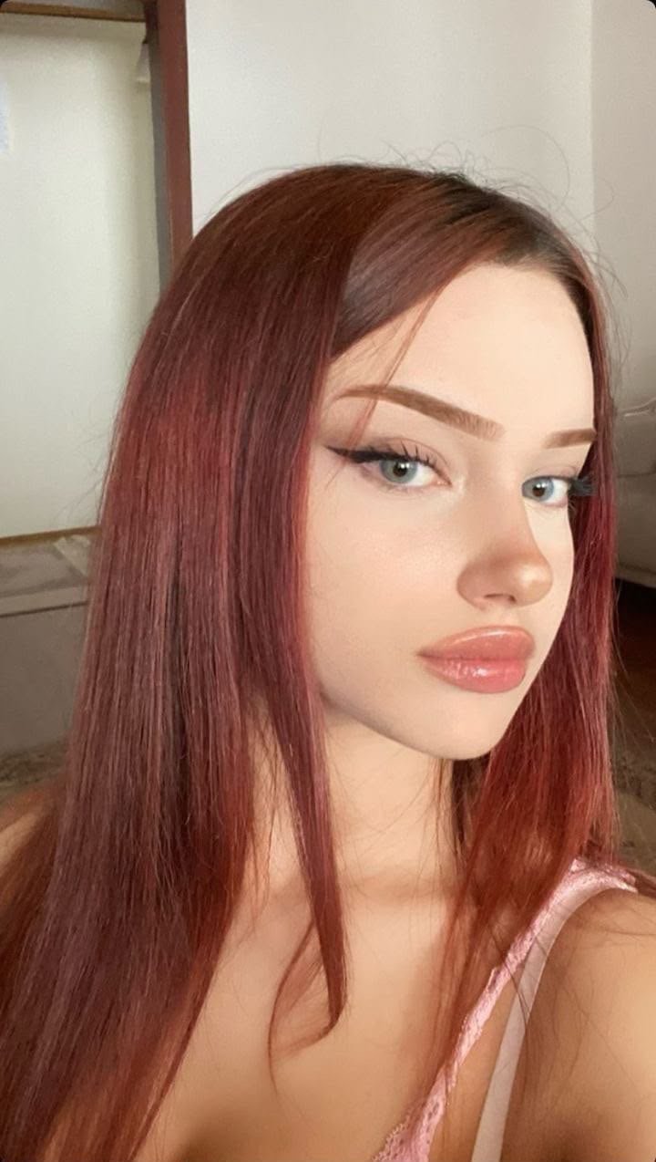 Close-up portrait of a young woman with long red hair, pink glossy lips, and winged eyeliner, looking slightly to the left.
