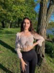 Young woman stands outdoors by a tree near a lake, wearing a floral off-the-shoulder crop top and black leggings.