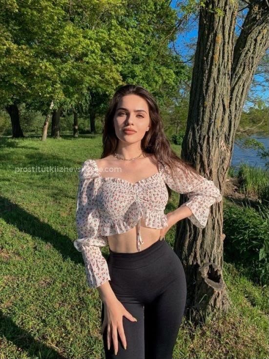Young woman stands outdoors by a tree near a lake, wearing a floral off-the-shoulder crop top and black leggings.
