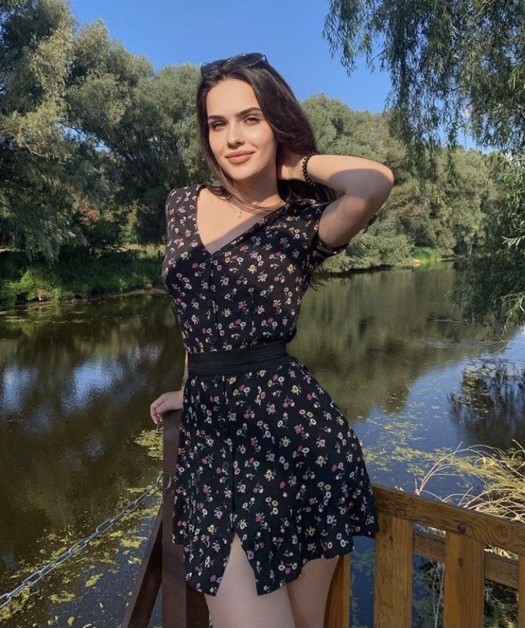 Young woman in a black floral dress stands on a wooden railing beside a calm lake on a sunny day.