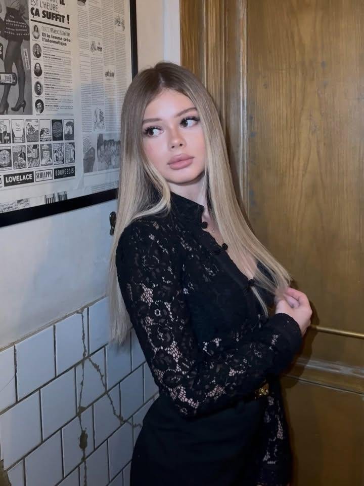 Young woman with long blonde hair in a black lace top standing beside a white tiled wall and wood door, looking to the side.