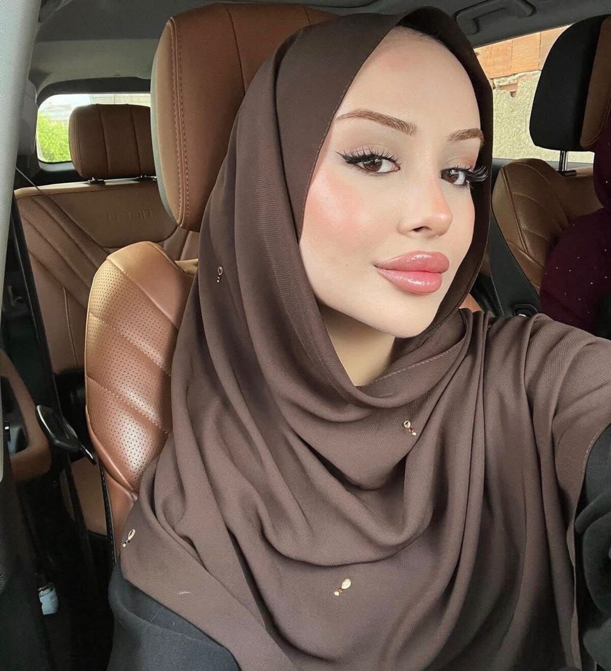 Close-up of a woman wearing a brown hijab inside a car, looking at the camera with soft makeup and pink lips.